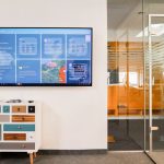 Sleek office interior featuring a wall-mounted TV screen and colorful dresser in a glass-walled room.