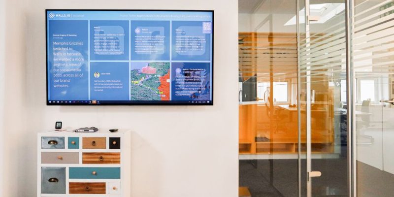 Sleek office interior featuring a wall-mounted TV screen and colorful dresser in a glass-walled room.