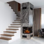 Interior of stylish living room with white stairway and wooden steps placed near marble column with burning fireplace at home