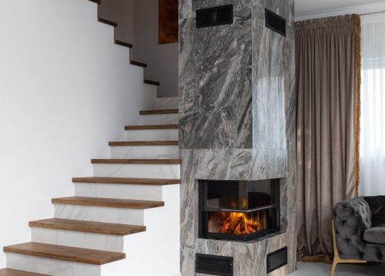Interior of stylish living room with white stairway and wooden steps placed near marble column with burning fireplace at home